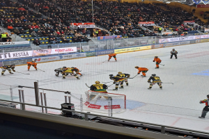 Eine Gruppe von Menschen, die Hockey auf einer Indoor-Eisfläche spielen, umgeben von einem Zaun, mit Zuschauern im Hintergrund und beleuchteter Stadionbeleuchtung darüber.
