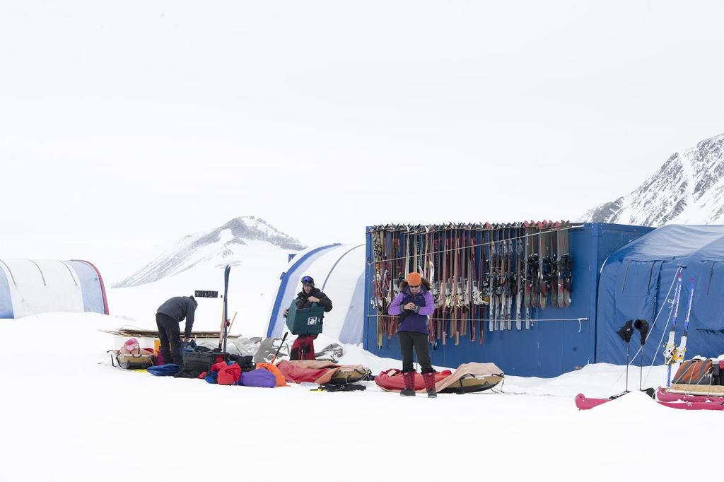 Drei Personen auf einer schneebedeckten Landschaft mit verstreuten Taschen, Zelte mit Skiern im Hintergrund und schneebedeckte Hügel unter einem klaren Himmel.
