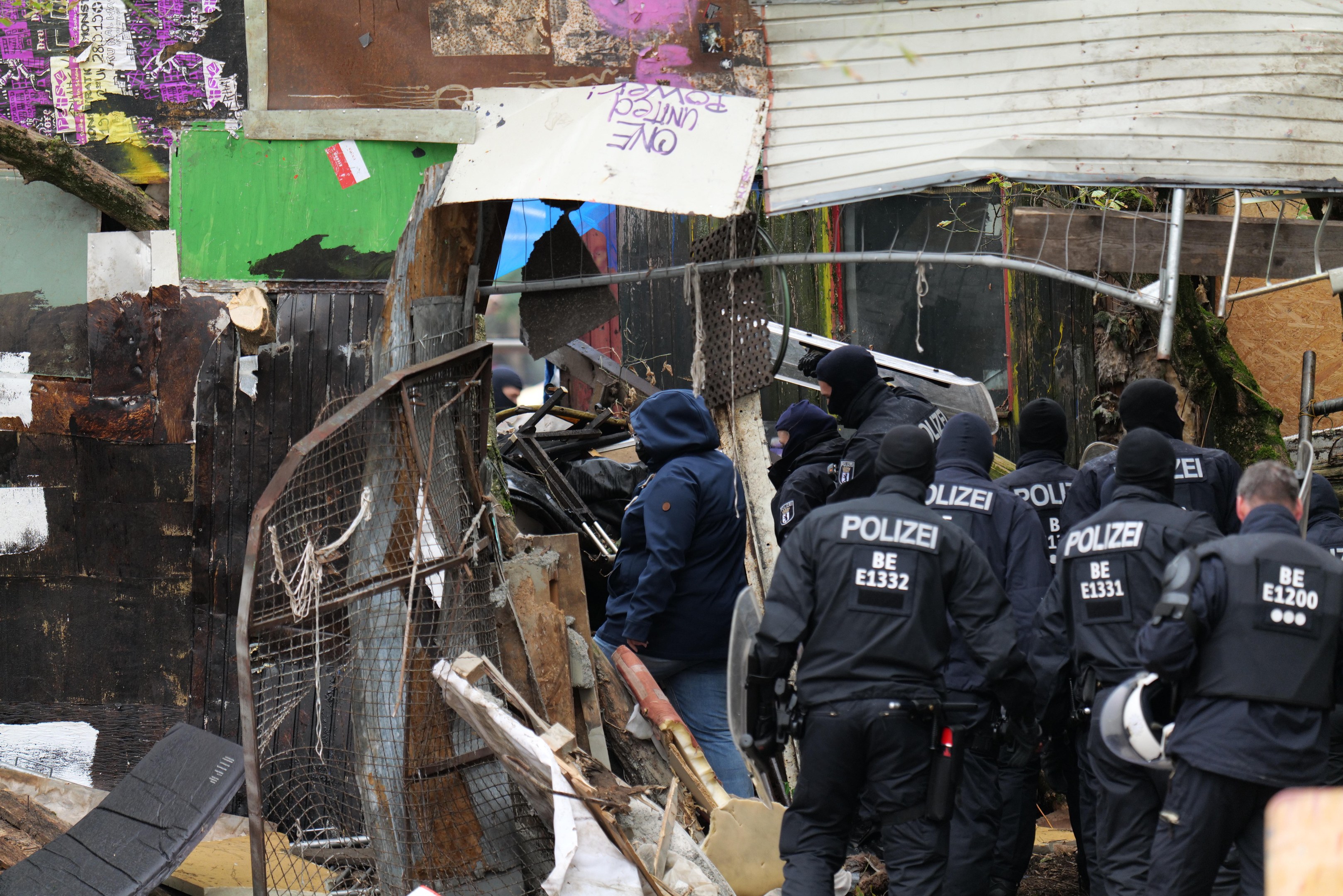 Group of police officers in black uniforms and helmets standing in front of a destroyed building surrounded by a metal fence, with scattered debris.