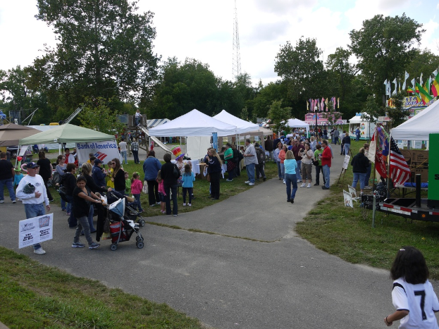 Eine Gruppe von Menschen, Stühle, Fahnen, Kinderstühle, Zelte und eine Tafel sind auf einer grünen Fläche mit Bäumen im Hintergrund aufgebaut.