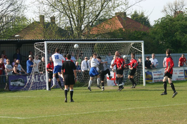 Fußballspieler auf einem Feld mit einem Tor und Zuschauern im Hintergrund, mit Bäumen und Häusern in der Ferne.