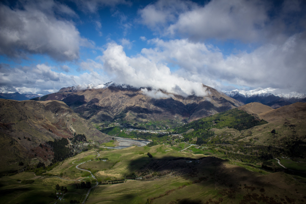 Eine Panoramansicht von einem Berg in Queenstown, Neuseeland, zeigt eine kurvenreiche Straße durch saftig grünes Gras und Bäume unter einem Himmel voller weißer, flauschiger Wolken.