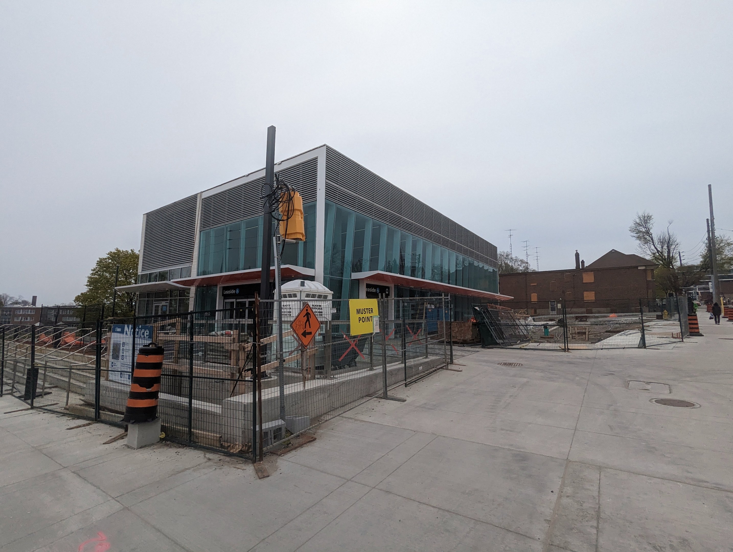 Large modern school building with many windows, surrounded by a metal fence, trees, signboards, and a few people and vehicles; cloudy sky above.