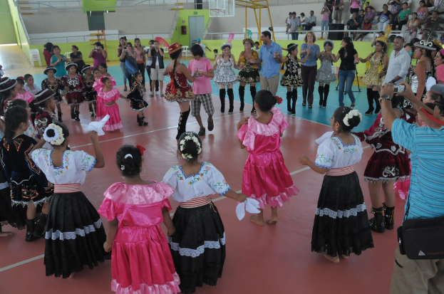 Eine Gruppe von Kindern in Kostümen, mit einem Mädchen und einem Jungen, die in der Mitte tanzen, während andere Menschen von oben rechts zuschauen.
