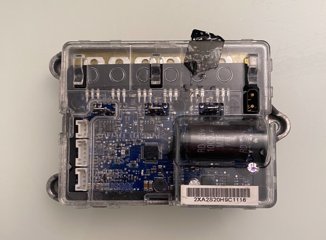 Close-up of a small electronic device in a plastic case on a white surface, showing a circuit board with a battery attached.