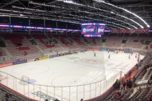 Eishockeyspiel in einer großen Arena zwischen den St. Louis Blues und Detroit Red Wings, mit Zuschauern auf den Sitzen und Stehplätzen in der Nähe der Eisfläche umgeben von einem Zaun und Metallrahmen, unter einem Dach mit Deckenleuchten und einem Anzeigebildschirm.