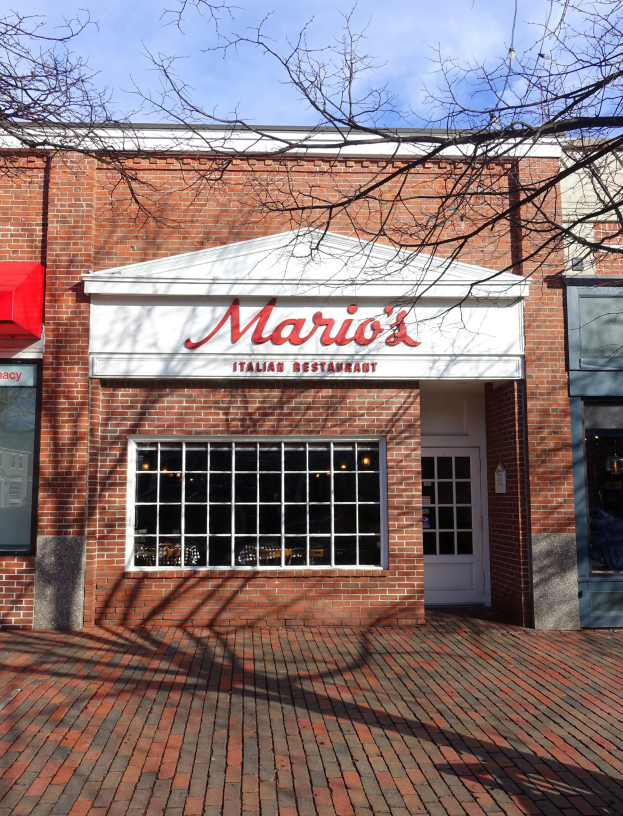 Mario's Italianisches Restaurant in St. Louis, Missouri, ein Gebäude mit Glasfenstern, einer Tür und einer Namensschild an der Wand, mit Zweigen im Vordergrund und einem Himmel voller weißer, flauschiger Wolken im Hintergrund.