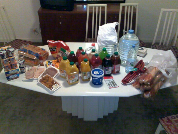 Ein Tisch mit einer Flasche, Essen und Anhängern darauf, umgeben von Stühlen, mit einem Fernseher in der Nähe.