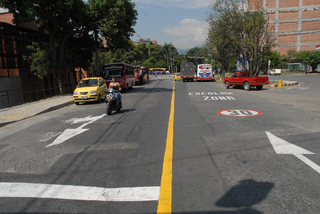 Eine Straßenszene mit mehreren Gebäuden, Bäumen, fahrenden Fahrzeugen, Schildern, Verkehrsampeln an Pfosten, einem blauen bewölkten Himmel und Warnschildern auf der Straße.