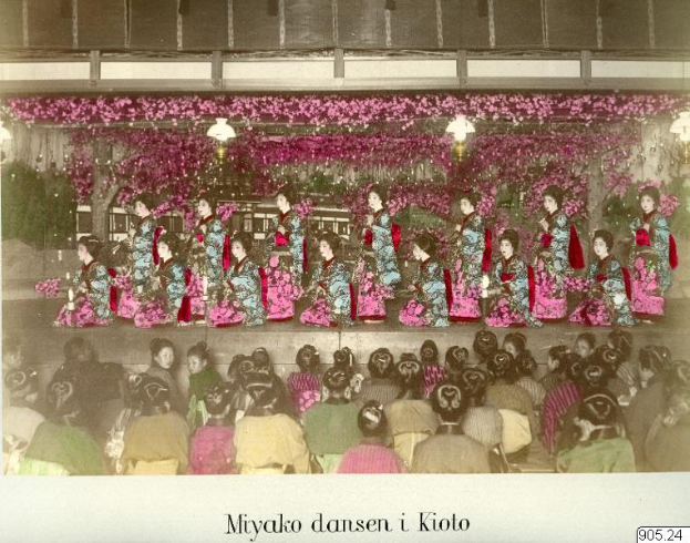 Ein altes Foto einer Gruppe von Frauen in bunten Kimonos, die auf einer Bühne mit Blumen und Lichtern sitzen, vor einem Publikum, das auf Stühlen sitzt, mit Text am unteren Rand des Bildes.