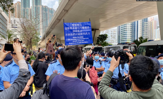 Eine Gruppe von Menschen vor einer Menschenmenge, einige mit Kameras und Handys, mit einem Banner mit der Aufschrift "Hong Kong Police Wants to Stop the Violence Against Women", Fahrzeuge, Bäume, Gebäude und ein bewölkter Himmel im Hintergrund.