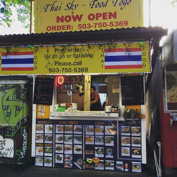 Ein Schaufenster mit verschiedenen Artikeln, darunter Getränkedosen, Speisekarten, eine Uhr, Gewürze in Plastikbehältern, Kondimente, Werbetafeln und Namensschilder, unter einem Himmel als Hintergrund.