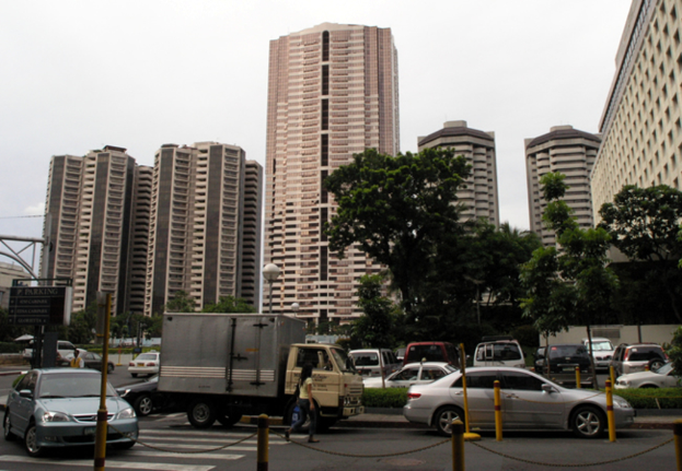 Eine Frau überquert eine Zebrastreifen, während Fahrzeuge auf der Straße fahren, mit Gebäuden, Bäumen und Pfählen im Hintergrund und einem an einem Pfahl befestigten Schild auf der linken Seite.