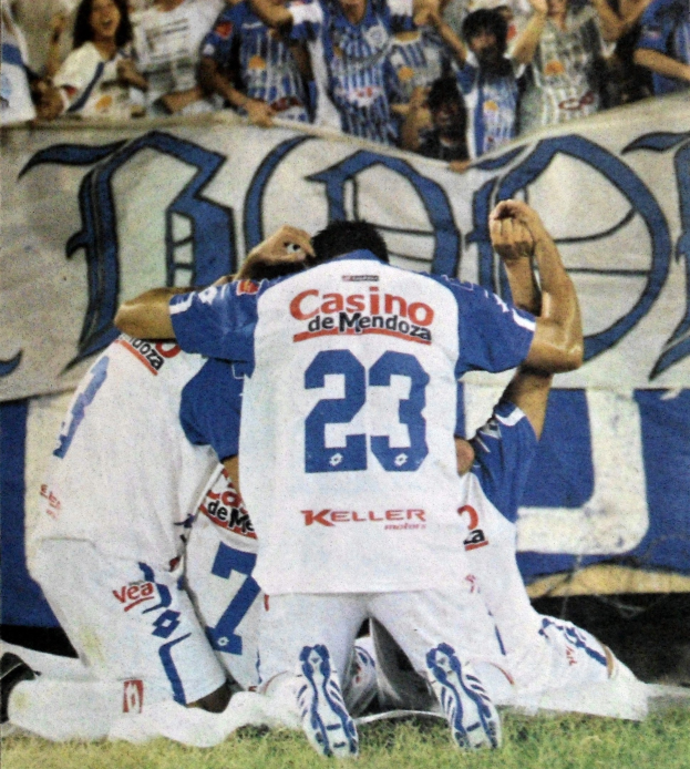 Gruppe von Fußballspielern feiert auf dem Boden mit einer jubelnden Menge und einem Banner im Hintergrund.