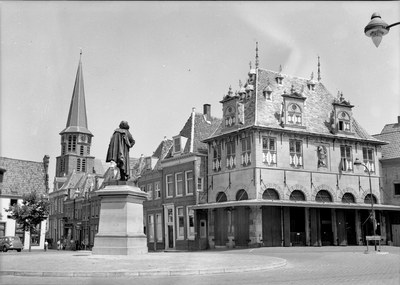Schwarzes und weißes Foto eines Stadtplatzes mit einer Statue auf einem Sockel in der Mitte, umgeben von Gebäuden, Straßenlaternen, Straßenlaternen, Fahrzeugen, Bäumen und einem klaren Himmel.