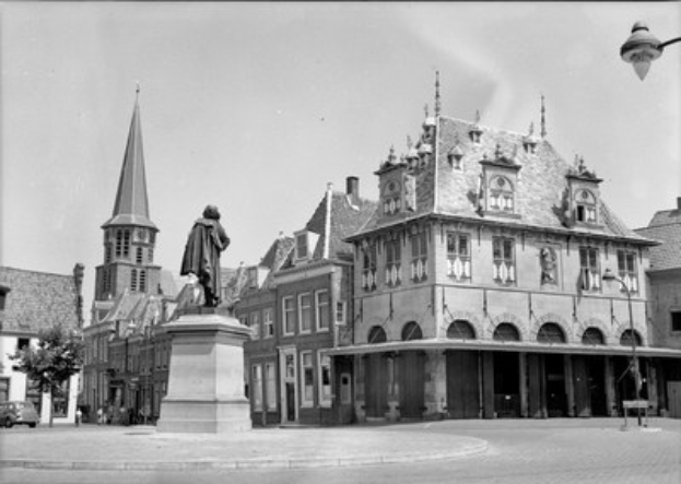 Schwarzes und weißes Foto eines Stadtplatzes mit einer Statue auf einem Sockel in der Mitte, umgeben von Gebäuden, Straßenlaternen, Straßenlaternen, Fahrzeugen, Bäumen und einem klaren Himmel.