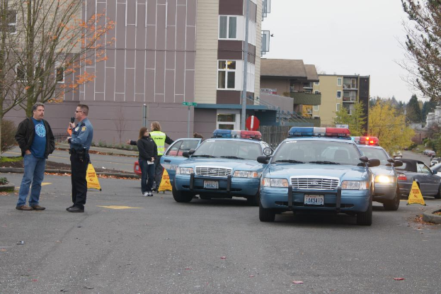 Autos auf einer Stra√üe mit vier Menschen in der N√§he, Geb√§ude mit Fenstern im Hintergrund, B√§ume und Warnkegel.