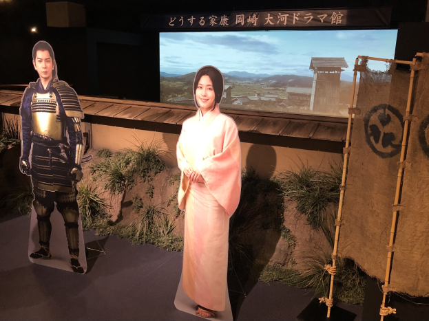 Zwei Menschen in traditioneller japanischer Kleidung – eine Frau in einem rosa Kimono und ein Mann in Samurai-Rüstung – lächeln gemeinsam auf einem Set mit Pflanzen, einem Bildschirm und Beleuchtungsausrüstung.