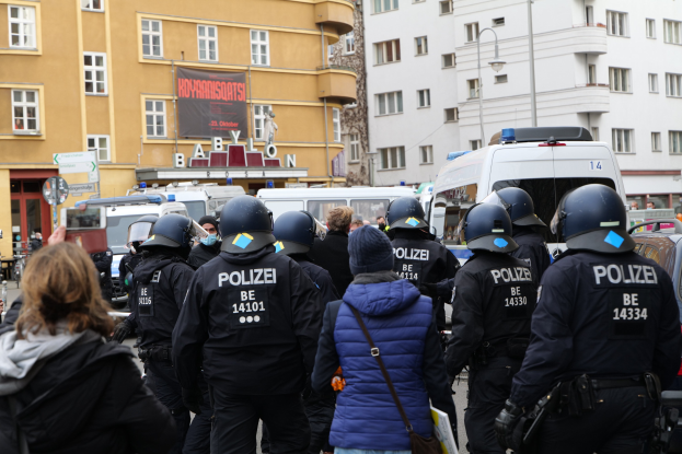 Polizeibeamte in Uniform vor einer Menge bei einer Demonstration in Berlin, mit Fahrzeugen, Gebäuden und einer Person mit einer Kamera im Bild.