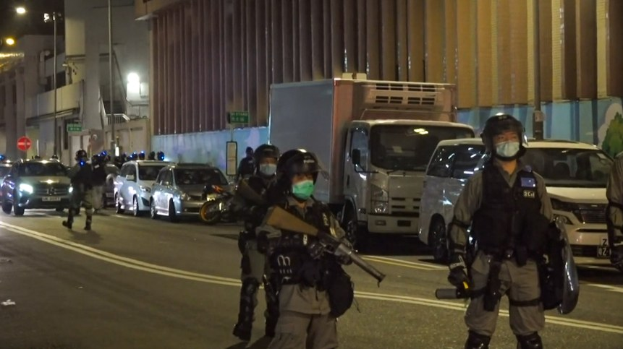 Eine Gruppe von Polizisten in Helmen, Handschuhen und Gesichtsmasken steht auf einer Straße bei Nacht, einige halten Waffen, mit Fahrzeugen, Laternenmasten, Schildern und Gebäuden im Hintergrund.