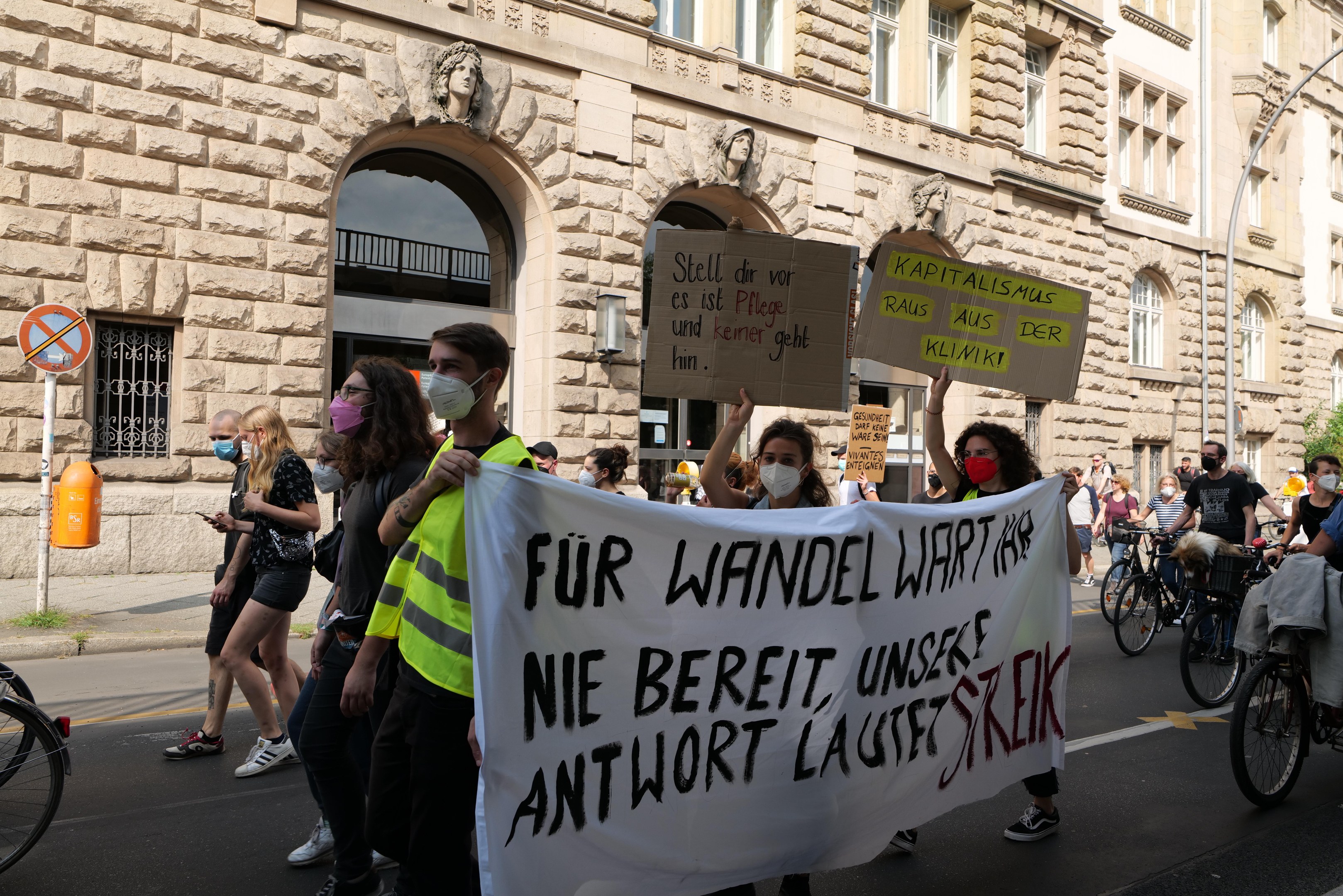 A group of people marches down a street, some holding protest signs and others riding bicycles, with a building featuring windows, arches, pillars, and sculptures in the background.