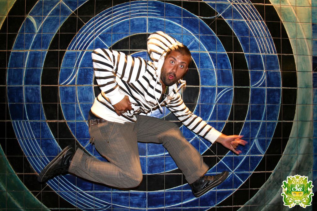 Ein Mann tanzt vor einem bunten Hintergrund und einem Logo mit Text in der rechten oberen Ecke.