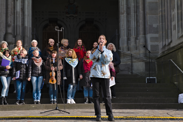 Eine Gruppe von Menschen, einige mit Musikinstrumenten, steht vor einem Gebäude mit Treppe und Geländer, während eine Frau in der Mitte in ein Mikrofon singt, umgeben von Mikrofonstativen.