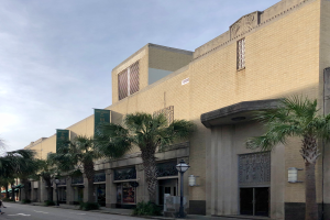 Das Hollywood Theatre in Hollywood, Kalifornien, ein großes Gebäude mit Palmen an seiner Seite, umgeben von Straßenmasten, Straßenlaternen, Kraftfahrzeugen auf der Straße, Fußgängern auf dem Gehweg, Büschen und einem bewölkten Himmel im Hintergrund.