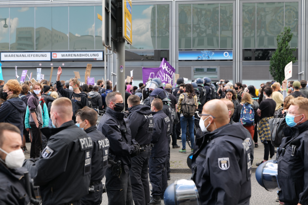 Eine große Gruppe von Menschen steht vor einem Gebäude, einige halten Schilder und tragen Helme, mit einem Pfahl und Schild im Vordergrund und einem Baum im Hintergrund, was auf eine Demonstration hindeutet.