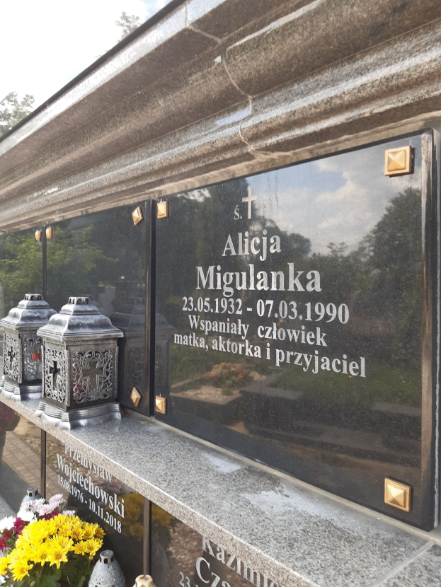 Denkmalstein mit einer Plakette, auf der 'St. Alicia Migulanka' steht, vor einem Gebäude, umgeben von Blumentöpfen und anderen Gegenständen, mit Bäumen und einem klaren blauen Himmel im Hintergrund.