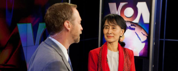 Ein Mann in einem grauen Blazer und weißem Hemd steht neben einer Frau in einem roten und weißen Kleid, beide vor einem Fernseher, der "Aung San Suu Kyi auf VOA" anzeigt, vor einem schwarzen Hintergrund.