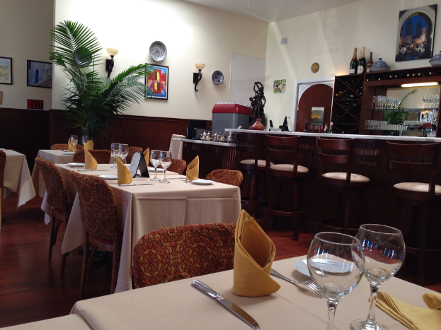 Ein Restaurantinnenraum mit Tischen und Stühlen, Gläsern, Servietten, Tellern und Löffeln auf den Tischen, einem langen Tisch mit Stühlen davor und Regalen mit Gläsern und Flaschen dahinter, einer Wand mit Fotorahmen, einer Uhr, zwei Lampen und einer Pflanze vor der Wand.