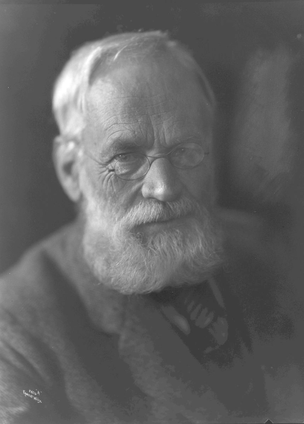 Ein älterer Mann mit Bart und Brille, der eine dunkle Hintergrund hat, hat einen ernsten Ausdruck und schaut konzentriert in die Ferne, mit ordentlich gekämmtem Haar und einem gut gepflegten Bart.