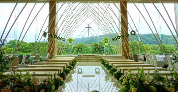 Eine Hochzeitszeremonie in einem großen Saal mit Tischen und Stühlen, die in einem Halbkreis angeordnet sind, dekoriert mit Blumenbouquets und anderen Gegenständen, vor einer Kulisse von Bäumen, Hügeln und einem klaren blauen Himmel.
