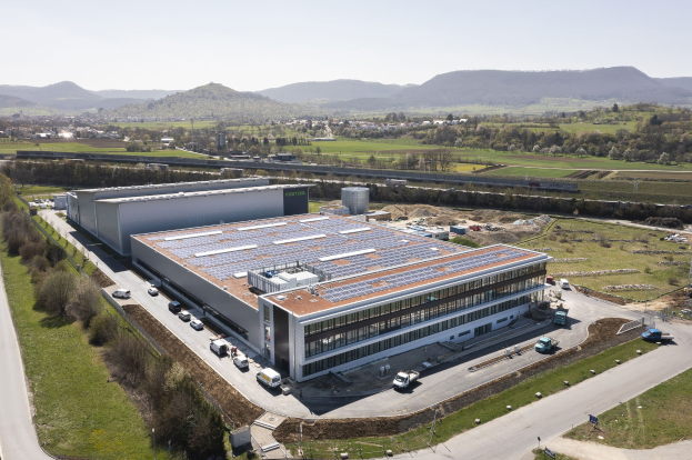Aerial view of a large building with solar panels on its roof, surrounded by vehicles, trees, grass, poles, and hills under a clear blue sky, identified as Germany's first solar-powered factory.