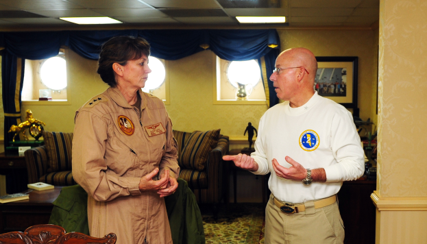 Ein Mann und eine Frau unterhalten sich in einem Wohnzimmer mit Möbeln, Büchern und einem Foto auf dem Regal, mit Fenstern und Lampen, die eine gemütliche Atmosphäre schaffen.