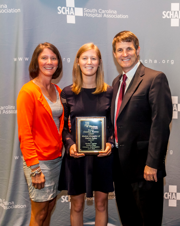 Drei Personen, zwei Frauen und ein Mann, stehen zusammen. Der Mann trägt einen schwarzen Anzug, eine Frau trägt ein orange-schwarzes Kleid und die andere Frau trägt ein schwarzes Kleid und hält eine Trophäe in der Hand. Sie stehen vor einem grauen Hintergrund.