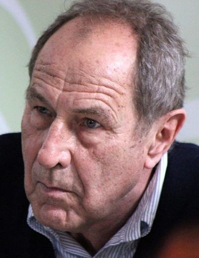 Ein Close-up eines Mannes in Anzug und Krawatte, der eine weiße Laborkittel und einen Stethoskop trägt und einen ernsten Gesichtsausdruck hat.