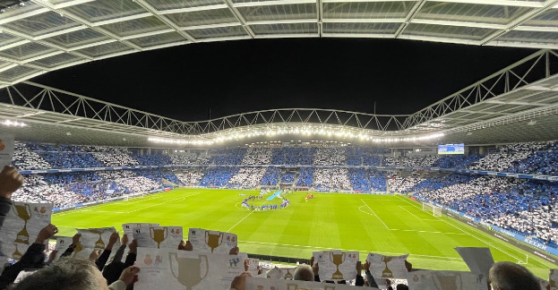 People holding signs and standing in front of a lit soccer stadium, with spectators visible in the background during a nighttime celebration.