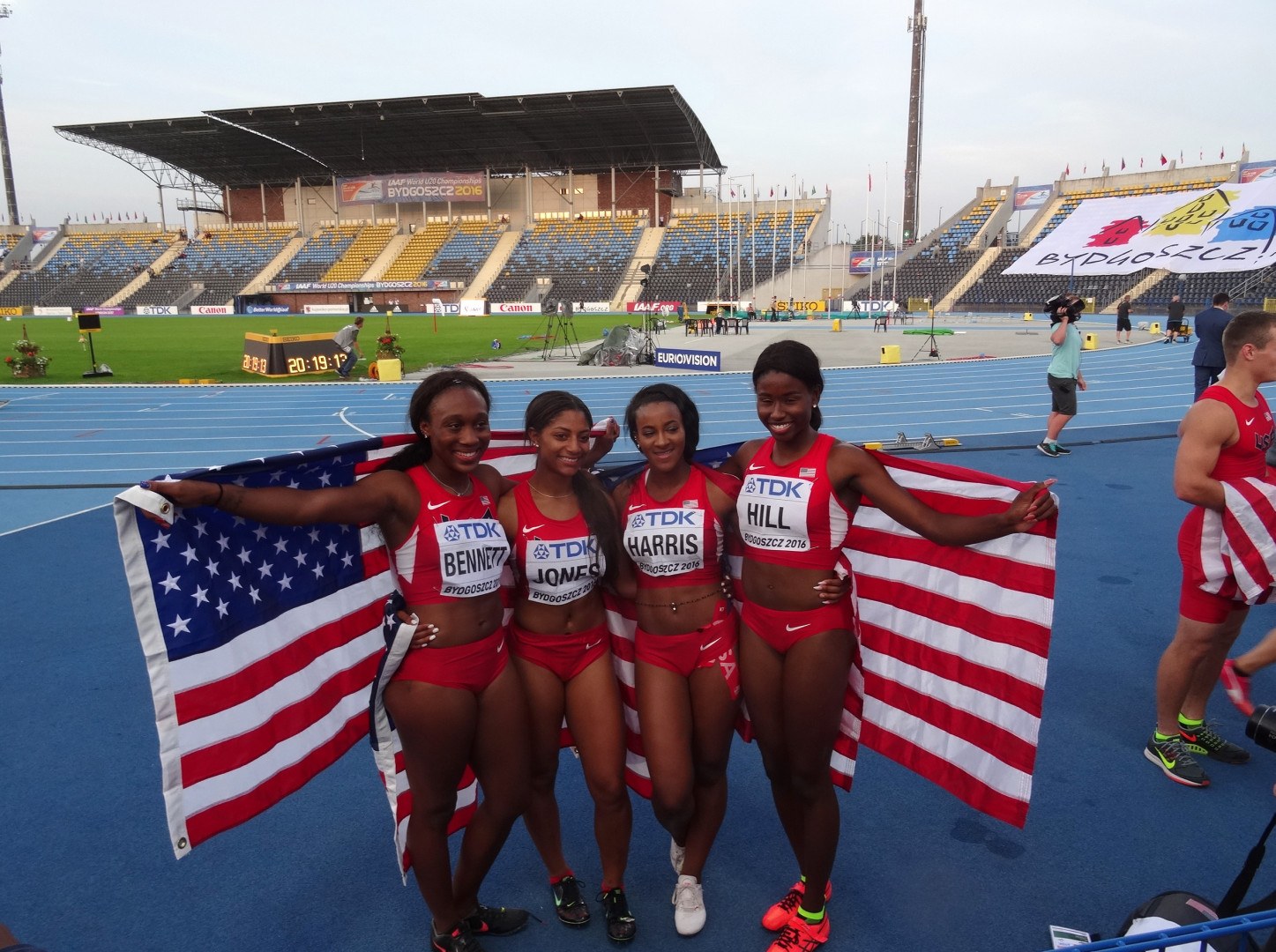 Vier Frauen In Sportbekleidung Stehen Auf Einer Laufbahn, Lachen Und Halten Amerikanische Flaggen, Mit Einer Stadion Und Zuschauern Im Hintergrund.
