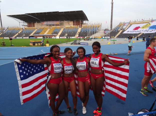 Vier Frauen In Sportbekleidung Stehen Auf Einer Laufbahn, Lachen Und Halten Amerikanische Flaggen, Mit Einer Stadion Und Zuschauern Im Hintergrund.