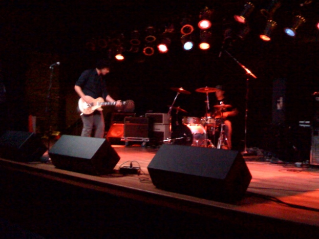 Zwei Personen, die auf einer Bühne Musikinstrumente spielen, mit Lautsprechern, anderen Instrumenten und Lampen an der Decke.