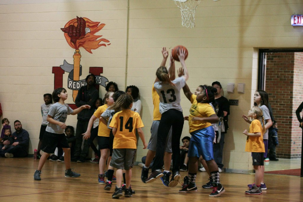 Kinder in gelben und grauen T-Shirts spielen Basketball, wobei ein Mädchen in einem grauen T-Shirt mit der Nummer 13 einen Wurf versucht. Im Hintergrund sitzt ein Mann in einem blauen Hemd neben einer weißen Wand mit Text, und rechts ist ein Ausgangsschild und ein rotes Backsteingebäude zu sehen.