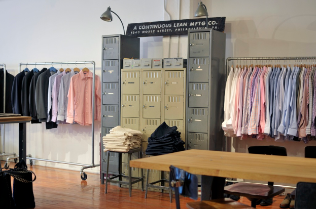 Ein Raum mit an einer Stange hängenden Hemden, Spinden, Kleidung auf Hockern, einem Boden und einer Wand mit Leuchten im Hintergrund.