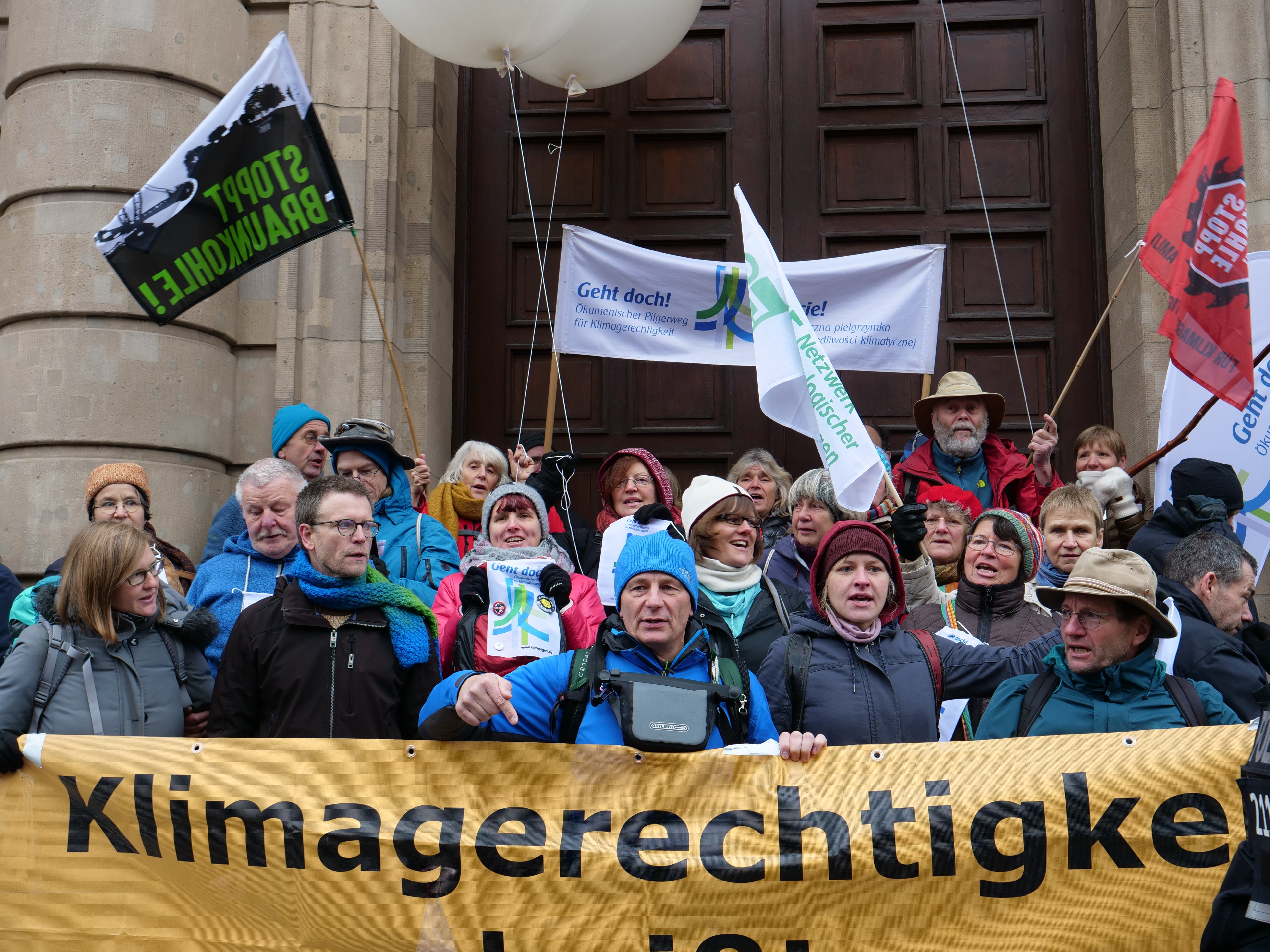 Eine Gruppe von Menschen steht vor einem Gebäude mit Säulen und einer Tür, hält ein Banner mit Text, Fahnen mit Stangen und Luftballons und nimmt an einem Klimastreik in Deutschland teil.
