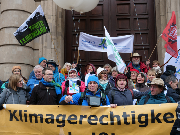 Eine Gruppe von Menschen steht vor einem Gebäude mit Säulen und einer Tür, hält ein Banner mit Text, Fahnen mit Stangen und Luftballons und nimmt an einem Klimastreik in Deutschland teil.