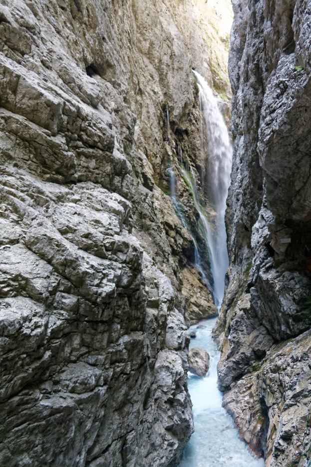 Kleiner Wasserfall, der über zerklüftete Felsen in einem steinigen Tal hinabstürzt, umgeben von grünen Hügeln unter strahlendem Sonnenlicht.