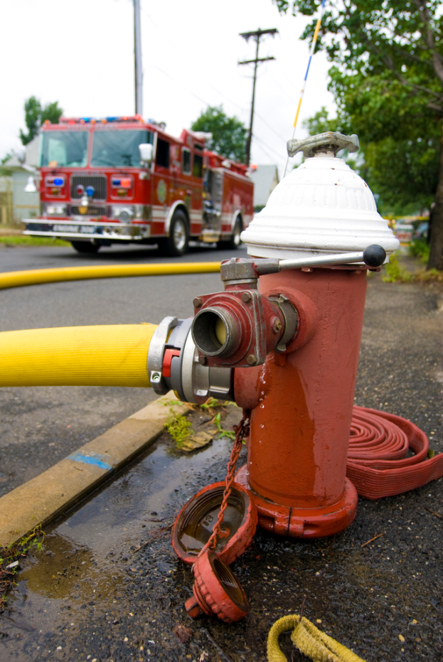 Ein Feuerhydrant und ein gelber Schlauch sind im Vordergrund zu sehen, im Hintergrund ein Bus, Stromleitungen, Masten und Bäume.