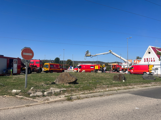 Eine Gruppe von Feuerwehrautos steht auf der Seite einer Straße, umgeben von Gras, Steinen, einem Schild, einem Haus, Pfählen, einem Kran, Drähten, Bäumen und einem bewölkten Himmel, wahrscheinlich nach einem Feuer an einer Tankstelle.