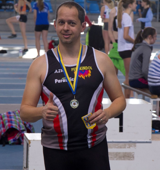 Ein Mann in schwarzem Trikot mit einer Medaille steht, während im Hintergrund einige Menschen trainieren.